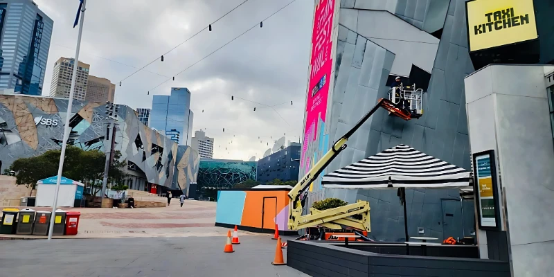 Maintenance technician on elevated work platform servicing a modern building facade in Melbourne's Federation Square with city skyline in background.